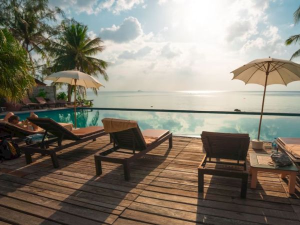 A serene poolside scene with lounge chairs, umbrellas, and palm trees overlooking a tranquil body of water under a partly cloudy sky.