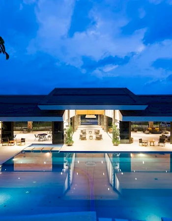 A modern building with an elegant outdoor pool area, illuminated at dusk, surrounded by lounge chairs and trees under a blue sky.