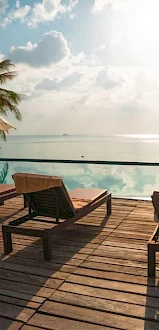 A serene poolside scene with lounge chairs, umbrellas, and palm trees overlooking a calm ocean under a partly cloudy sky at sunset.