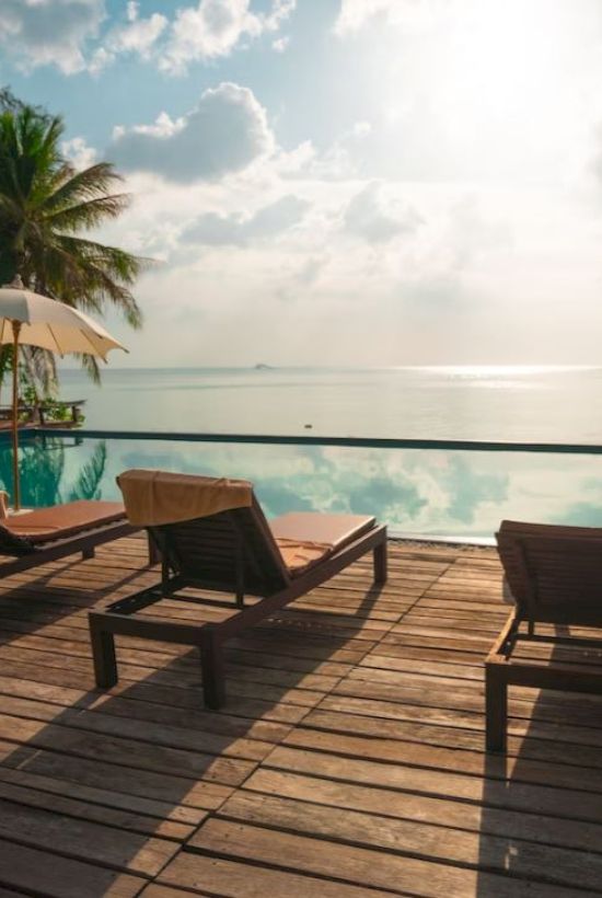 A serene beachside pool area with lounge chairs, umbrellas, palm trees, and ocean view on a sunny day.
