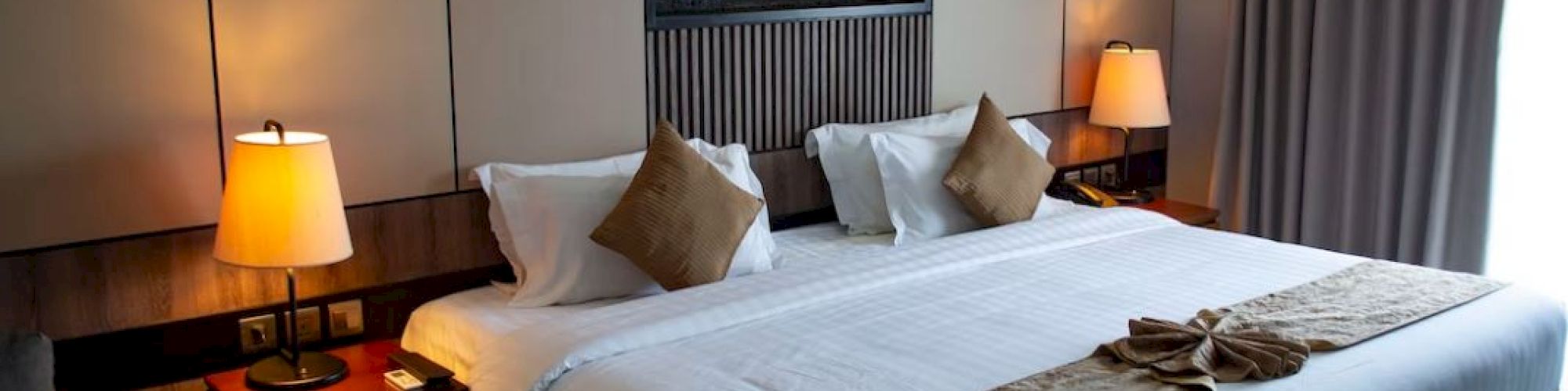 A cozy hotel room features a large bed with beige and brown accents, two lamps on bedside tables, and a large window with curtains.