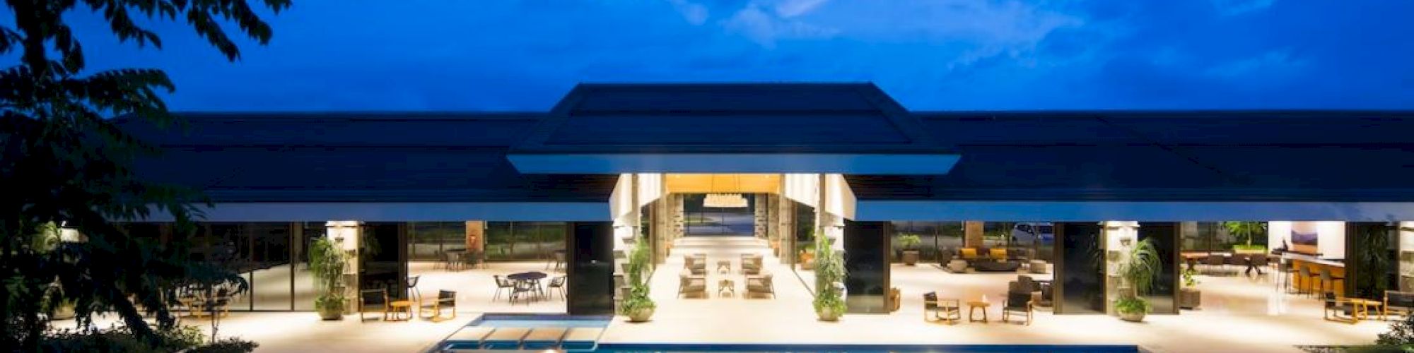 A luxurious resort at dusk with a large, illuminated swimming pool, surrounded by seating areas and lush greenery, under a clear blue sky.