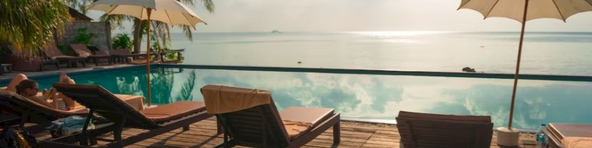 A serene poolside view with lounge chairs, umbrellas, palm trees, and a stunning ocean backdrop, all under a bright, partly cloudy sky.