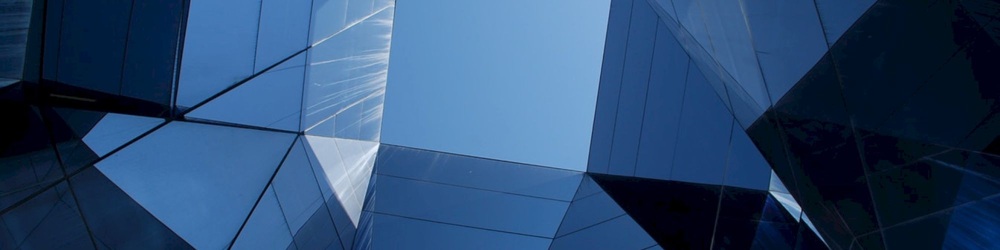 The image shows a modern architectural structure with mirrored glass surfaces reflecting the sky, creating a fragmented and geometric visual effect.