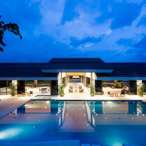 A luxurious modern building with a large illuminated pool in front, surrounded by outdoor seating, under a twilight sky.
