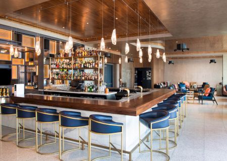 The image shows a modern bar with a large, fully stocked counter, blue bar stools, and hanging pendant lights in a stylishly designed interior.