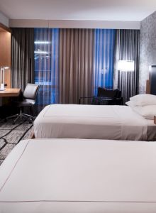 The image shows a modern hotel room with two neatly made beds, a wall-mounted TV, a desk with a chair, and large windows with curtains.