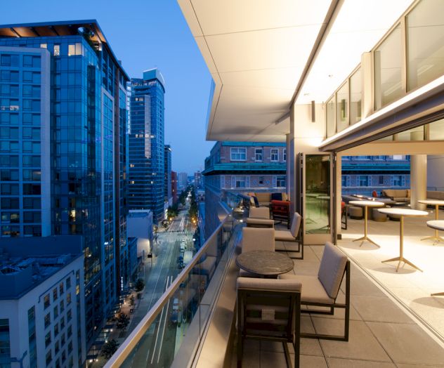 A rooftop terrace with tables and chairs overlooks a cityscape with modern buildings and a street below, illuminated by evening lights.
