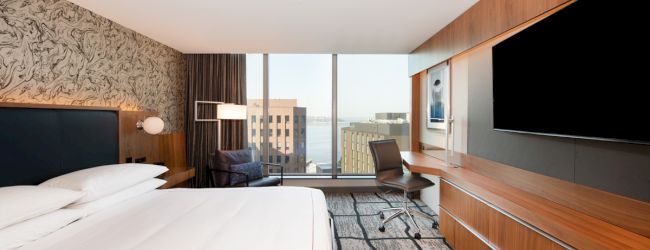 This image shows a modern hotel room with a bed, desk, chair, large TV, and a window view of buildings and water in the distance.