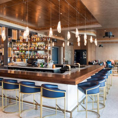 A modern bar with blue barstools, pendant lighting, and a well-stocked assortment of bottles. The space includes a seating area with lounge chairs.