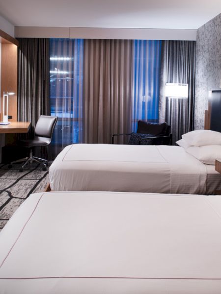 The image shows a modern hotel room with two neatly made beds, a flat-screen TV, a desk with a chair, and contemporary decor and lighting.