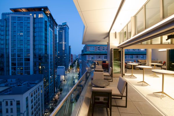 An evening cityscape view from a modern balcony, featuring outdoor seating and tables, with tall buildings lining a well-lit street below.