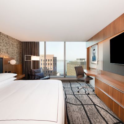 This image shows a modern hotel room with a large bed, a desk, a chair, a TV, and a window with a city view.