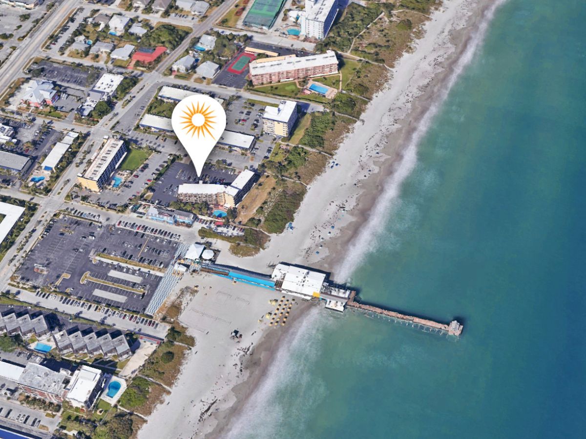 An aerial view of a coastal area with buildings, a beach, and a pier extending into the ocean. A location pin with a sun symbol is also present.