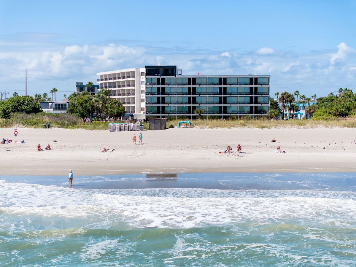 A beachfront scene shows a modern building, sandy shore, people relaxing, and waves hitting the shoreline under a blue sky.