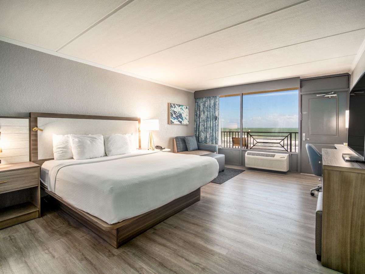 This image shows a modern hotel room with a large bed, a desk with a TV, a sofa, and a sliding door leading to a balcony with an ocean view.