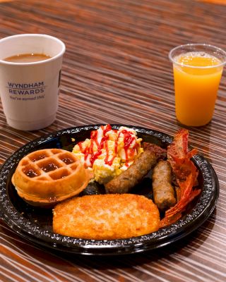 The image shows a breakfast plate with a waffle, scrambled eggs with ketchup, sausage, bacon, and a hash brown, accompanied by coffee and orange juice.