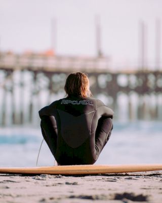 A person in a wetsuit sits on the sand beside a surfboard, facing the ocean with a pier in the background.