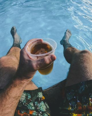 A person is sitting by a pool with their feet in the water, holding a plastic cup with a drink, and wearing patterned swim trunks.