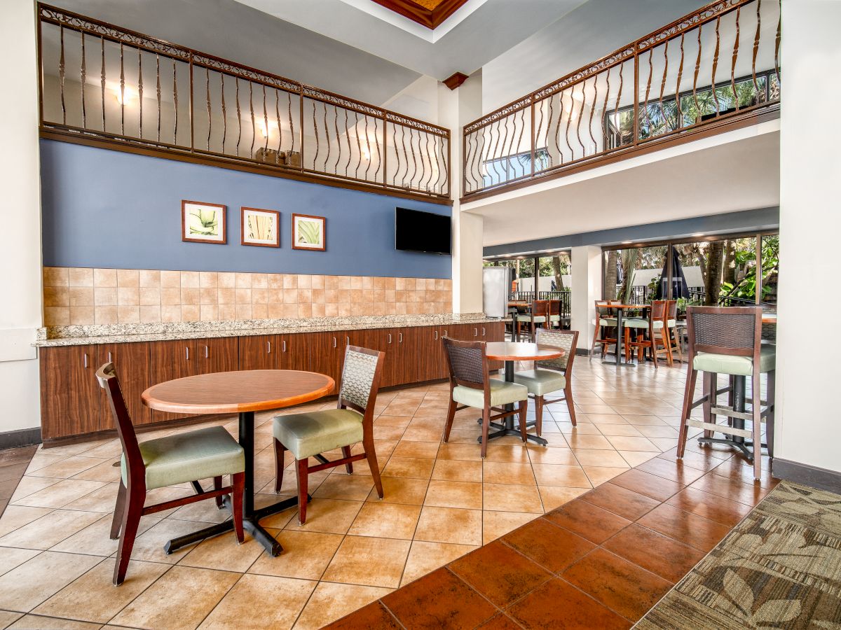 The image depicts an indoor dining area with round tables, chairs, and a bar counter under a double-height ceiling.