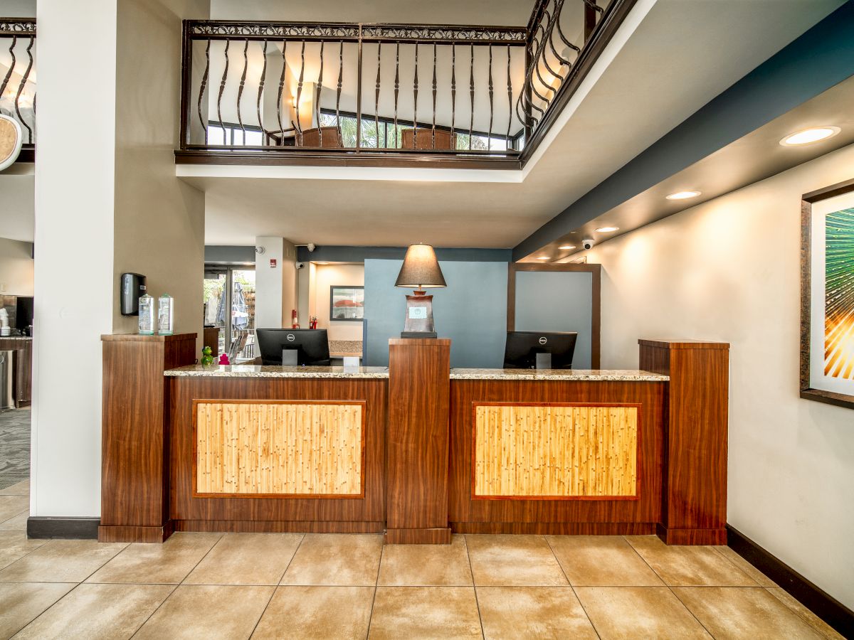 A hotel lobby with a front desk, two computer monitors, a lamp, and artwork on the wall. There are railings leading to an upper level.