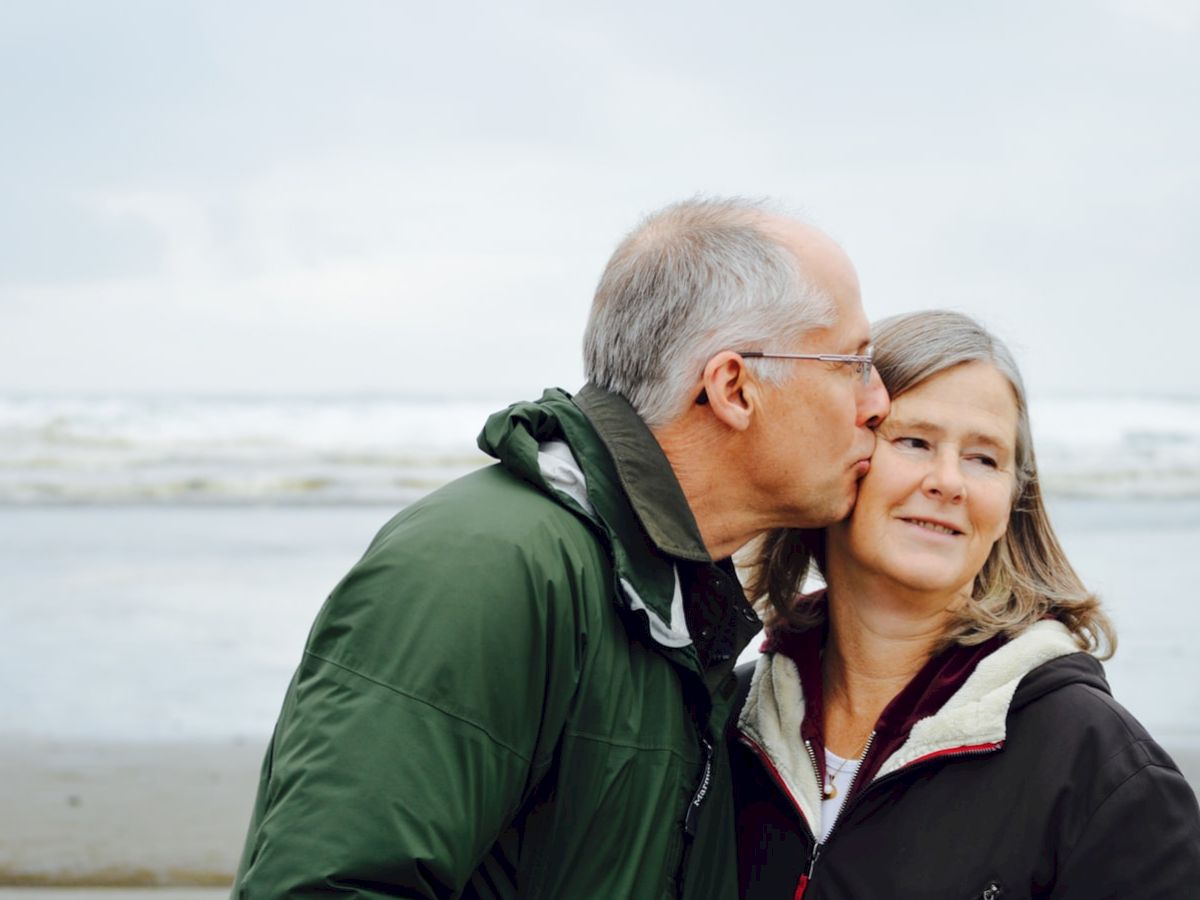 A man in a green jacket is kissing a woman's cheek while standing on a beach with waves in the background.