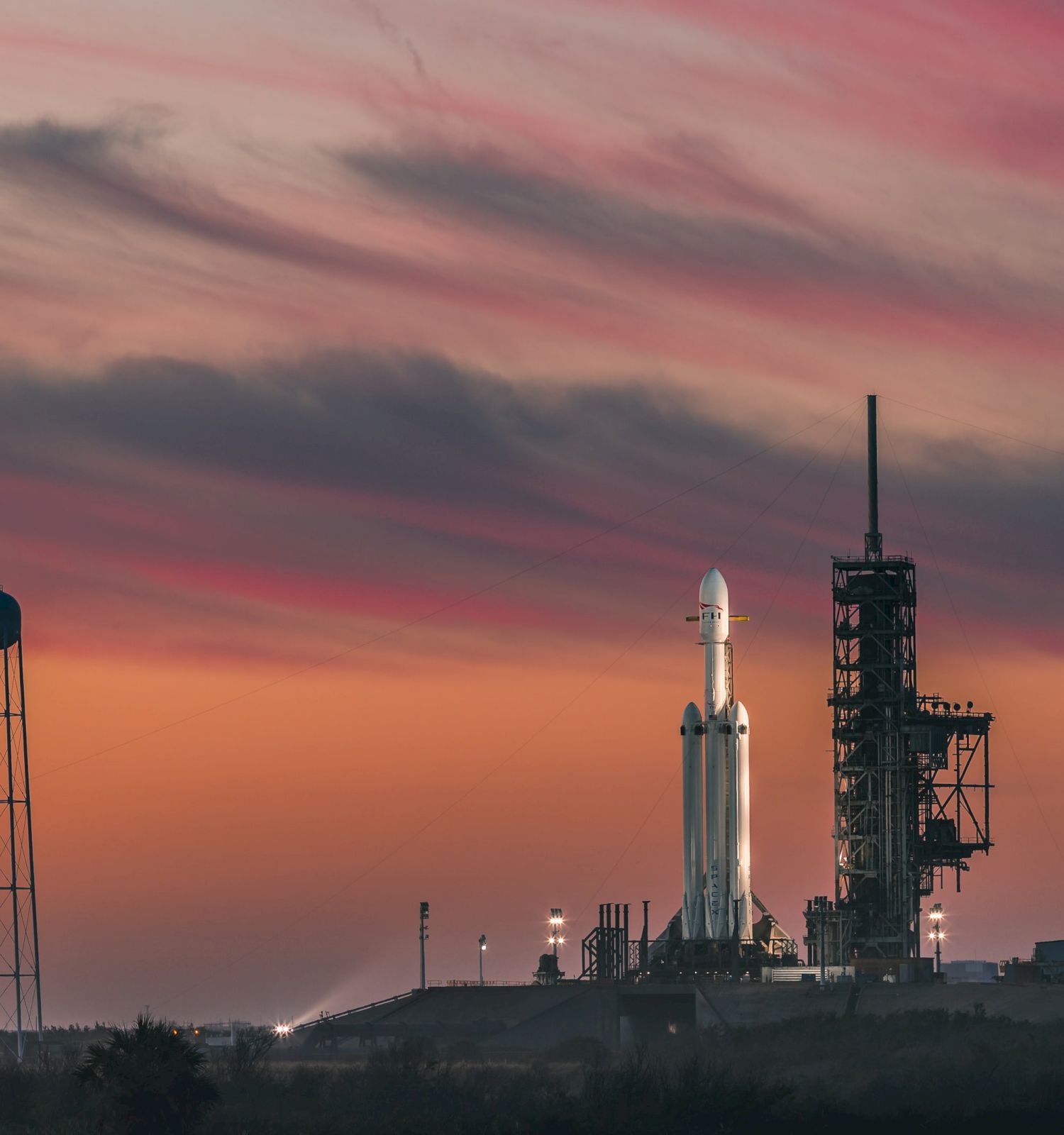 A rocket on a launch pad is silhouetted against a colorful sunset sky, with a water tower and structures nearby.