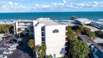 The image shows a coastal hotel with a parking lot, nearby buildings, and a pier extending into the ocean, under a partly cloudy sky.