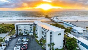 A beachfront hotel with a parking lot is shown at sunset, overlooking the ocean and a pier extending into the water.