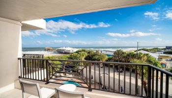 A balcony overlooking a scenic coastal view with a railing, two chairs, and a small table, facing the ocean. A beautiful sunny day.