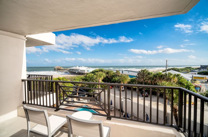 A balcony overlooking a scenic coastal view with a railing, two chairs, and a small table, facing the ocean. A beautiful sunny day.