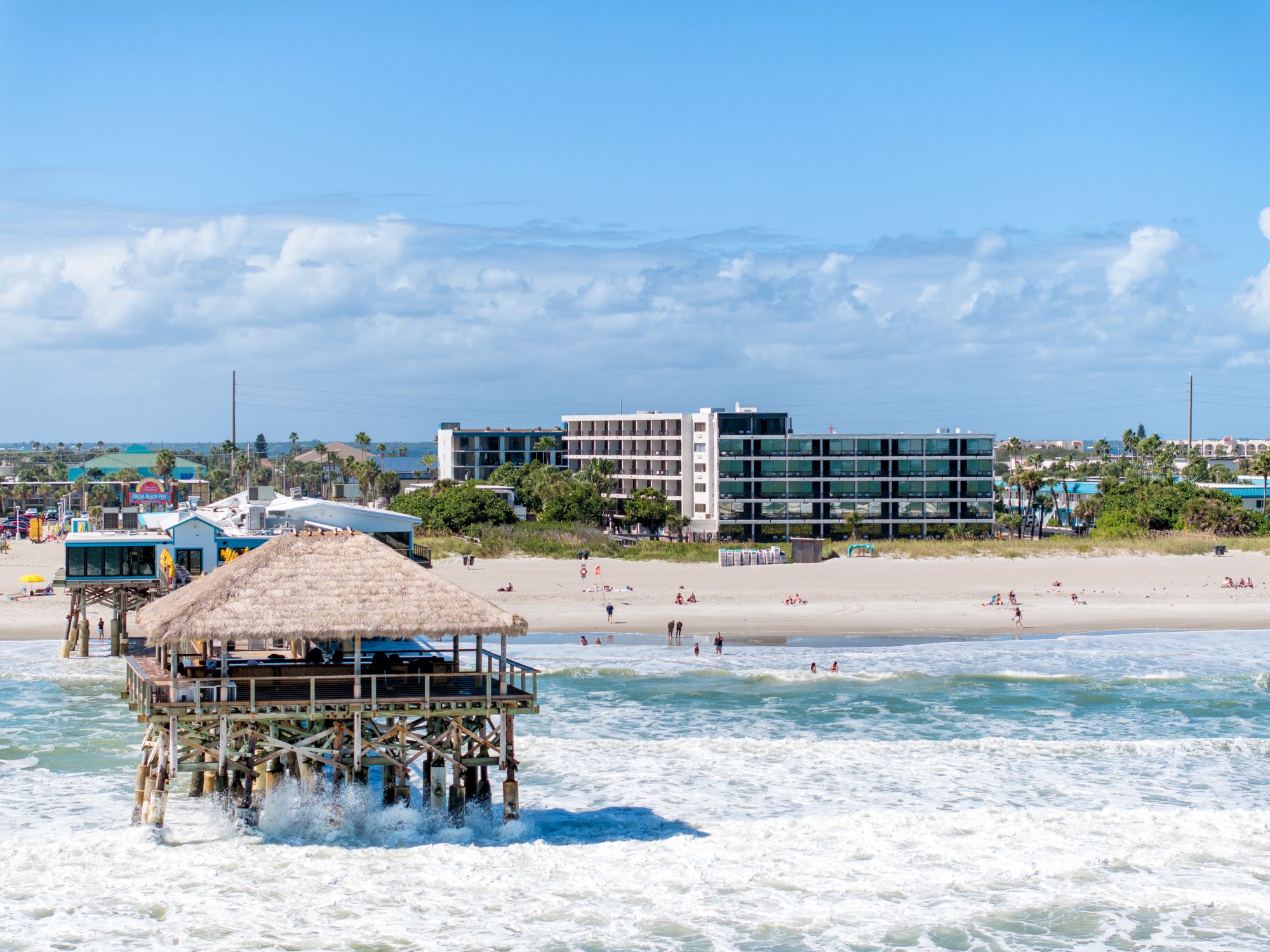 A beach scene with several people, buildings, and a thatched-roof structure on stilts in the water, under a partly cloudy blue sky.