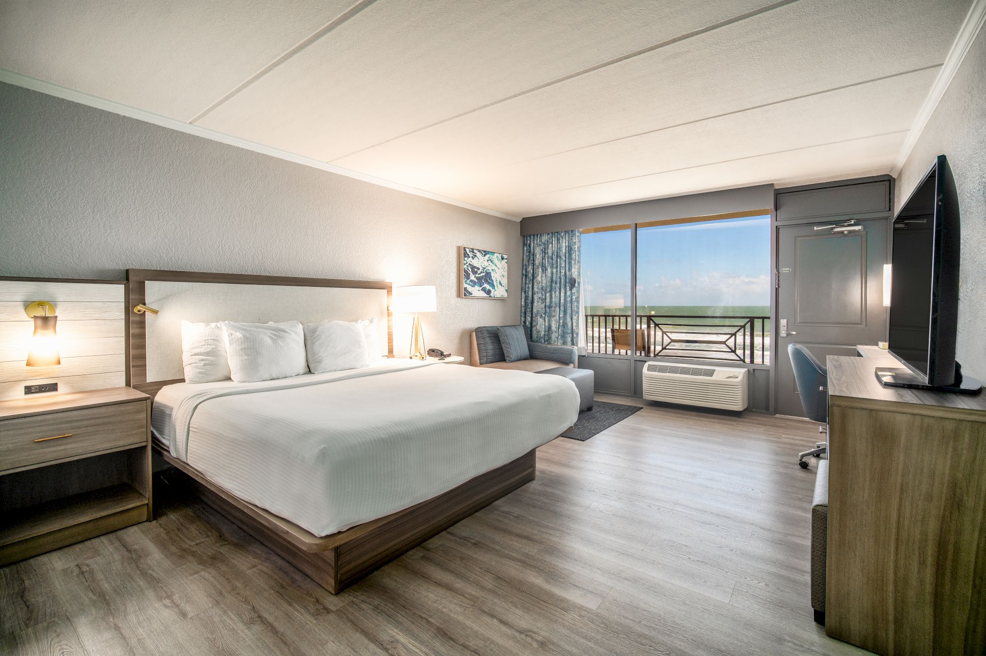 A modern hotel room with a king-sized bed, nightstands, a sofa, a desk, a TV, and a large window with a view of the ocean.