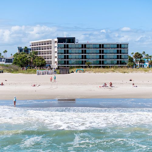 A beach scene with people on the shore, calm waves, and a modern building with many windows in the background, surrounded by greenery.