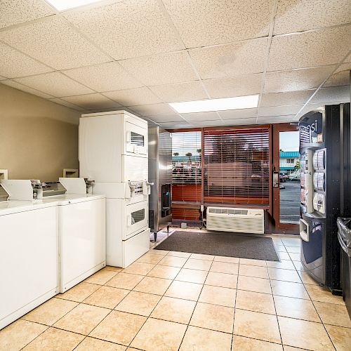 A small laundry room with washing machines, dryers, tiled floor, vending machines, and a window with blinds. A door leads outside.