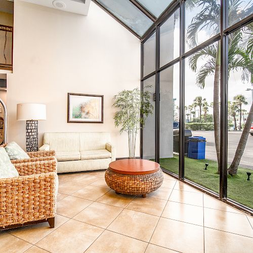 A bright, modern sitting area with wicker furniture, a round coffee table, a floor lamp, and large windows looking out to palm trees and a parking lot.