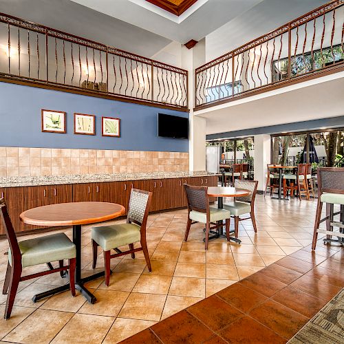 The image shows a modern dining area with wooden tables and chairs, tile flooring, framed pictures, and an upstairs balcony with metal railings.