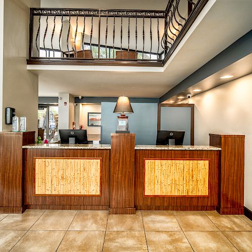 A hotel reception area with a wooden front desk, two computers, a lamp, and a hallway leading to other areas. There is art on the wall.