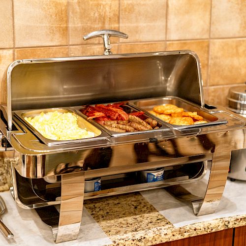 The image shows a buffet setup with various breakfast foods including scrambled eggs, bacon, sausages, and hash browns in a metal warmer.