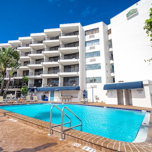 An outdoor pool area with lounge chairs is in front of a multi-story white building with balconies under a bright blue sky.