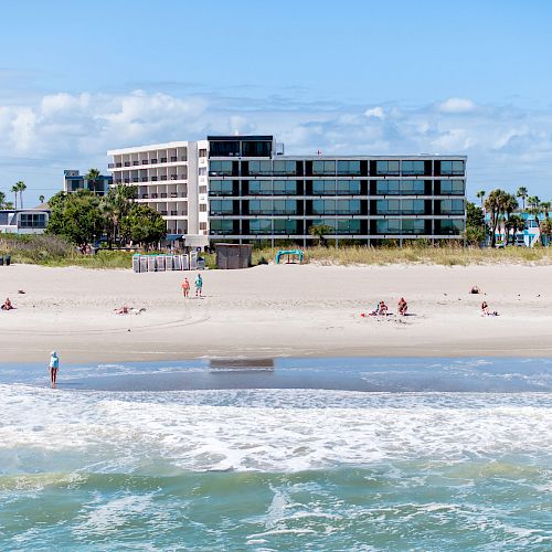 A beach scene with several people relaxing on the sand and a large building in the background with blue skies and some clouds.