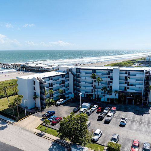 A beachfront hotel with a parking lot, adjacent to a sandy beach and a pier extending into the ocean, under a clear blue sky.