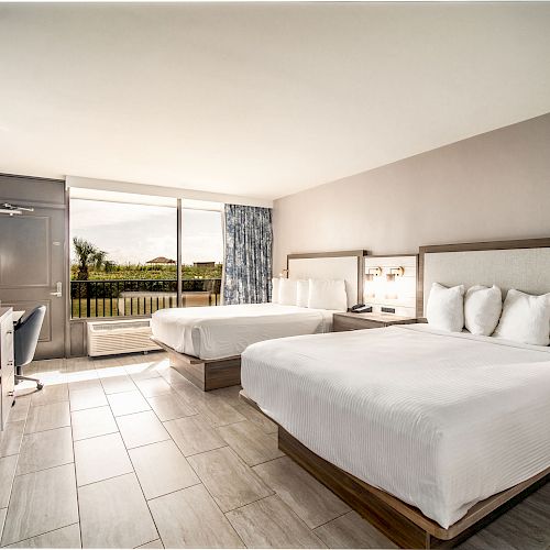 The image shows a modern hotel room with two double beds, a flat-screen TV, a desk, a chair, and a large window offering a view of greenery outside.