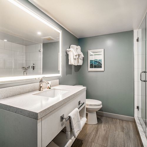 A modern bathroom with a large mirror, sink, toilet, shelves, towels, framed artwork, gray walls, and a glass-enclosed shower.