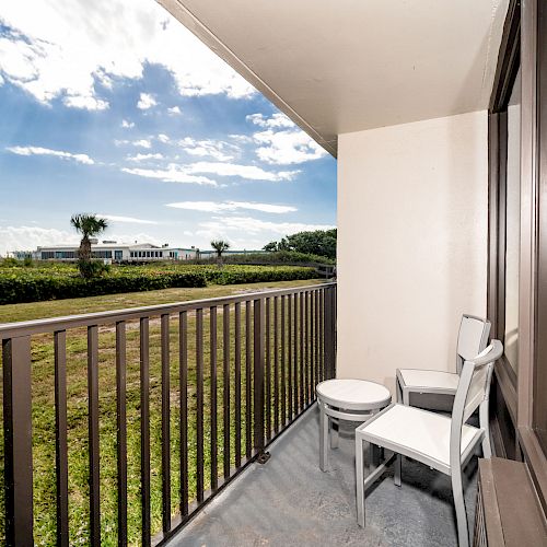 A balcony with two white chairs and a small table overlooks a grassy area and trees under a bright blue sky with scattered clouds.