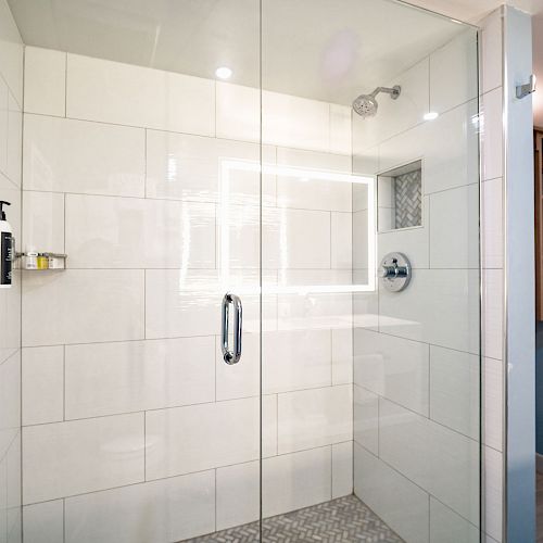 A modern shower with glass doors, white tiles, and a built-in shelf, located in a bathroom with light blue walls and shelving.