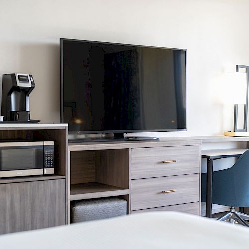 A modern hotel room setup with a TV, coffee maker, microwave, desk lamp, and a blue desk chair.