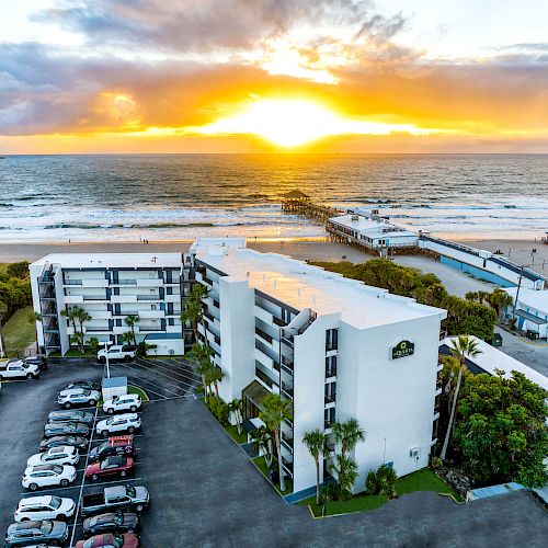 A coastal resort with parked cars, multi-story buildings, a sandy beach, and a pier extending into the ocean, all under a vibrant sunset sky.