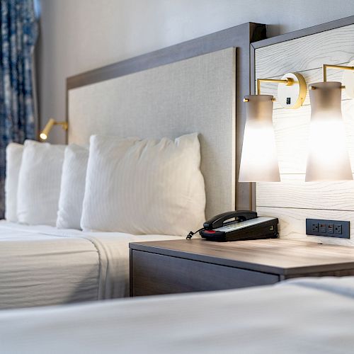 The image shows a hotel room with two beds, a nightstand with a telephone and power outlets, and two wall-mounted lights above the headboard.