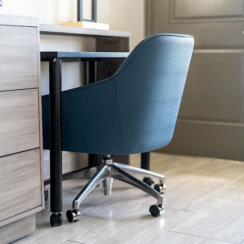 The image shows a home office setup with a blue swivel chair, wooden drawers with brass handles, a desk, and a door in the background.